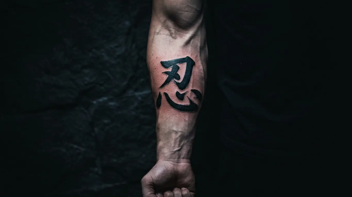 Un primer plano de un antebrazo con un tatuaje de caligrafía negra fresca del kanji de perseverancia 忍, que representa una hoja sobre un corazón.