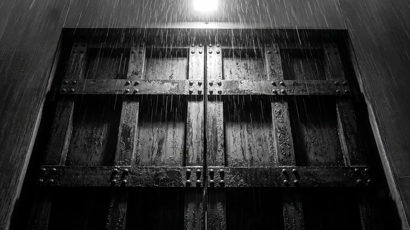 Plano medio contrapicado de una gran puerta metálica industrial oxidada y cerrada en un callejón nocturno y lluvioso de Tokio.